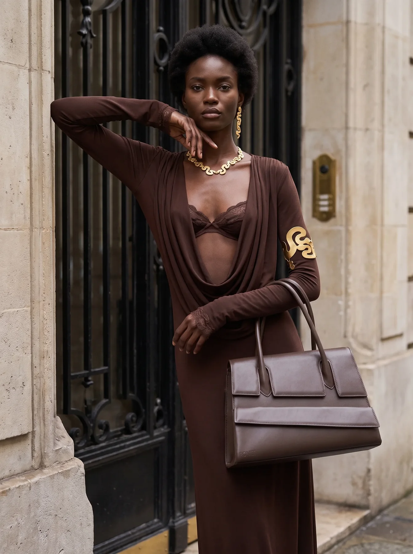 Chocolate draped gown, gold jewelry and structured bag against a Paris doorway